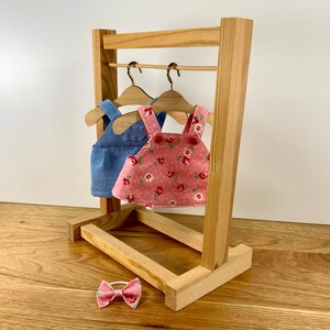 K&ouml;nnte beinhalten: Ein h&ouml;lzerner Kleiderst&auml;nder f&uuml;r Puppen mit zwei Kleiderb&uuml;geln, an denen ein rosa Kleid mit Blumenmuster und eine blaue Jeans-Latzhose h&auml;ngen. Im Vordergrund befindet sich eine rosafarbene Schleife mit wei&szlig;em Zentrum.