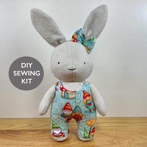 May include: A plush rabbit toy with light grey fur, long ears, and a small bow on its head. The rabbit wears light blue overalls featuring a colourful pattern of gnomes, mushrooms, and flowers. A circular grey badge displays the text DIY SEWING KIT.