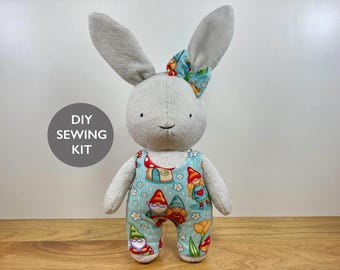 May include: A plush rabbit toy with light gray fur, long ears, and a small bow on its head. The rabbit wears light blue overalls featuring a colorful pattern of gnomes, mushrooms, and flowers. A circular gray badge displays the text DIY SEWING KIT.