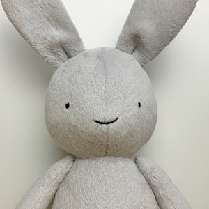 May include: A grey plush bunny with a simple black embroidered smile. The bunny has long, floppy ears and a soft, fuzzy texture.