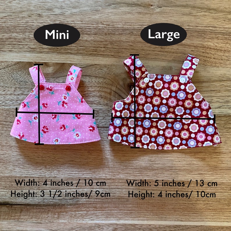 K&ouml;nnte beinhalten: Zwei Puppenkleider, eines rosa mit wei&szlig;en Punkten und eines rot mit wei&szlig;en und blauen Punkten. Das rosa Kleid ist mit "Mini" beschriftet und misst 10 cm breit und 9 cm hoch. Das rote Kleid ist mit "Large" beschriftet und misst 13 cm breit und 10 cm hoch.