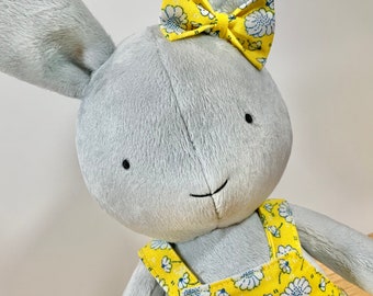May include: A gray plush bunny with long ears and a yellow floral bow. The bunny is wearing a yellow and white floral overall.