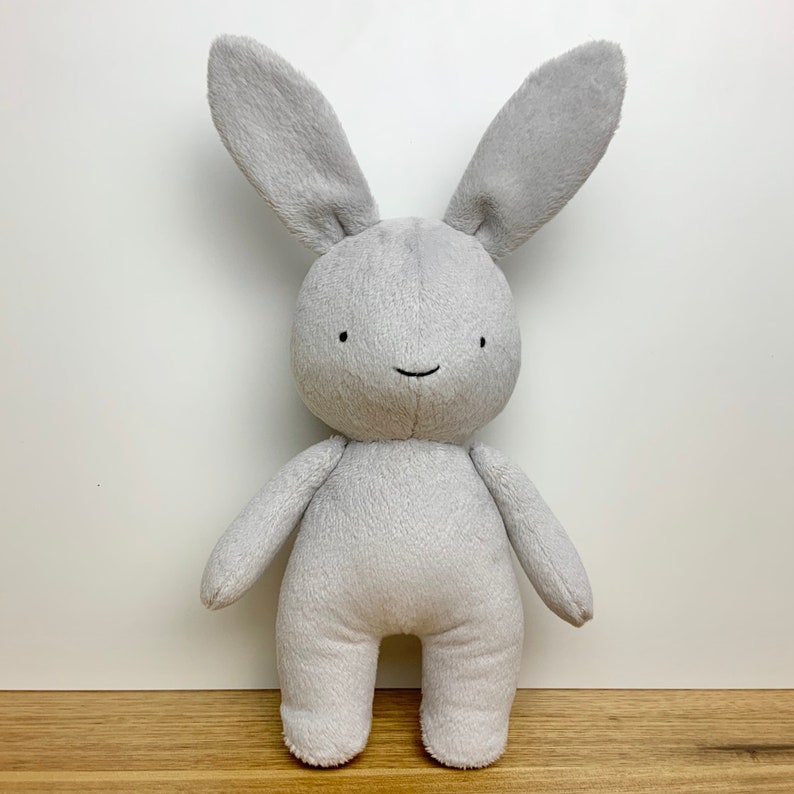 May include: A grey plush bunny toy with a simple, smiling face. The bunny has long ears and a soft, fluffy texture.