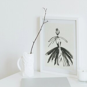 May include: A white framed print of a black and white illustration of a woman wearing a dress. The print is on a white table with a white vase and a twig, and a white candle in a glass jar.