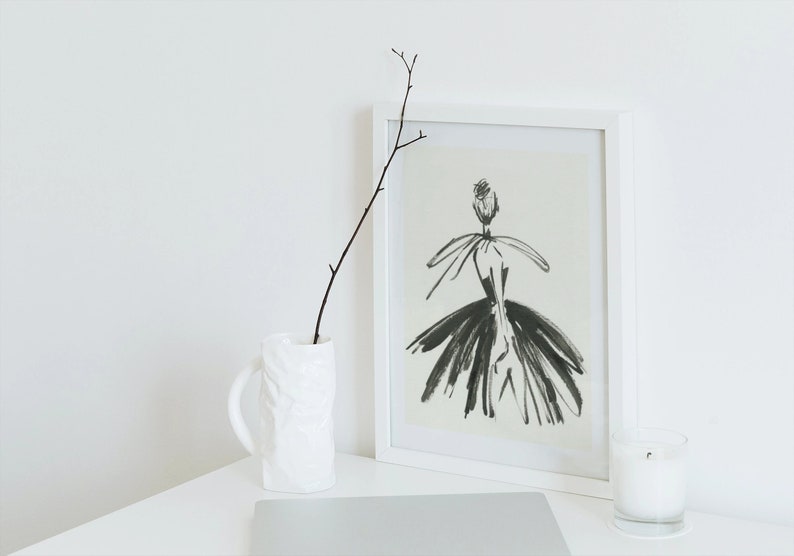 May include: A white framed print of a black and white illustration of a woman wearing a dress. The print is on a white table with a white vase and a twig, and a white candle in a glass jar.