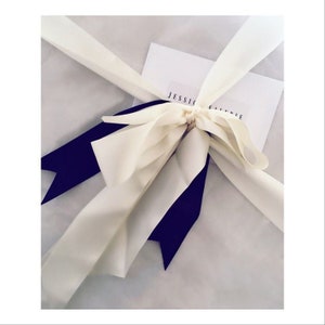 May include: A white gift bag with a white ribbon tied in a bow with two black ribbon tails. The bag has a white card with the text "JESSIC" on it.
