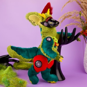 May include: A plush creature with green and teal fur, a black visor-like face, yellow horns, and a red mohawk. It features red hip patches with spiderweb icons, yellow claws, and large ears with Roman numeral markings, standing on a black hand-shaped stand.