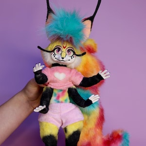 May include: A colourful plush toy with a yellow and black body, a rainbow tail, and a turquoise mohawk. The toy has a pink heart on its chest and black eyes with pink accents.