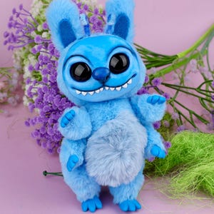 May include: A plush blue creature with large black eyes, a wide grin, and fluffy ears. The toy has a light blue belly and blue feet and hands. It is posed in front of a bouquet of purple and white flowers.