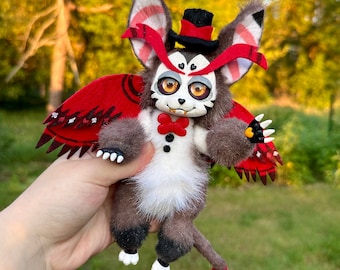 May include: A plush toy with a gray and white fur, red wings, and a red tail. The toy is wearing a black top hat and a red bow tie. The toy has large, yellow eyes and a toothy grin.