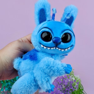 May include: A plush blue creature with large black eyes, a wide grin, and fluffy fur. The toy has large ears and a small antenna. The creature is held in a hand against a purple background, with some flowers in the bottom right corner.
