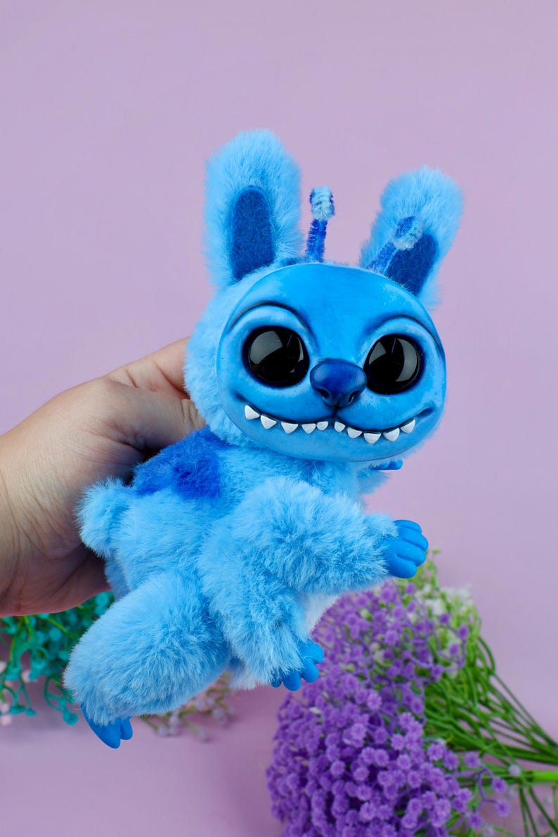 May include: A plush blue creature with large black eyes, a wide grin, and fluffy fur. The toy has large ears and a small antenna. The creature is held in a hand against a purple background, with some flowers in the bottom right corner.