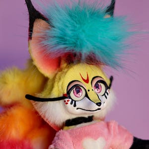 Op de afbeelding: Een kleurrijk pluche speelgoed met een geel gezicht, roze ogen en een zwarte neus. Het speelgoed heeft een blauw en groene mohawk, een roze shirt met een wit hart en een regenboogkleurige staart.