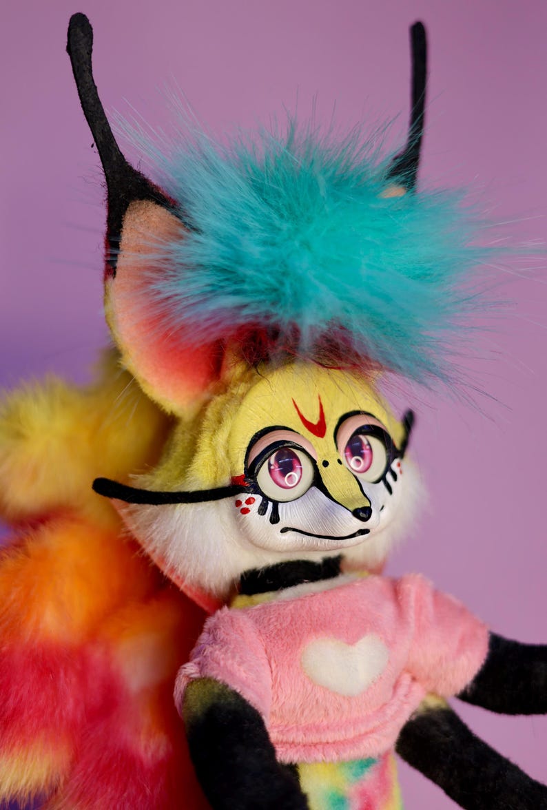 Op de afbeelding: Een kleurrijk pluche speelgoed met een geel gezicht, roze ogen en een zwarte neus. Het speelgoed heeft een blauw en groene mohawk, een roze shirt met een wit hart en een regenboogkleurige staart.