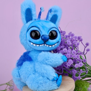 May include: A plush blue creature with large black eyes, a wide grin, and bunny-like ears. The stuffed animal is sitting on a small wooden stool, with a bouquet of purple flowers in the background. The toy has a patch of darker blue on its back.