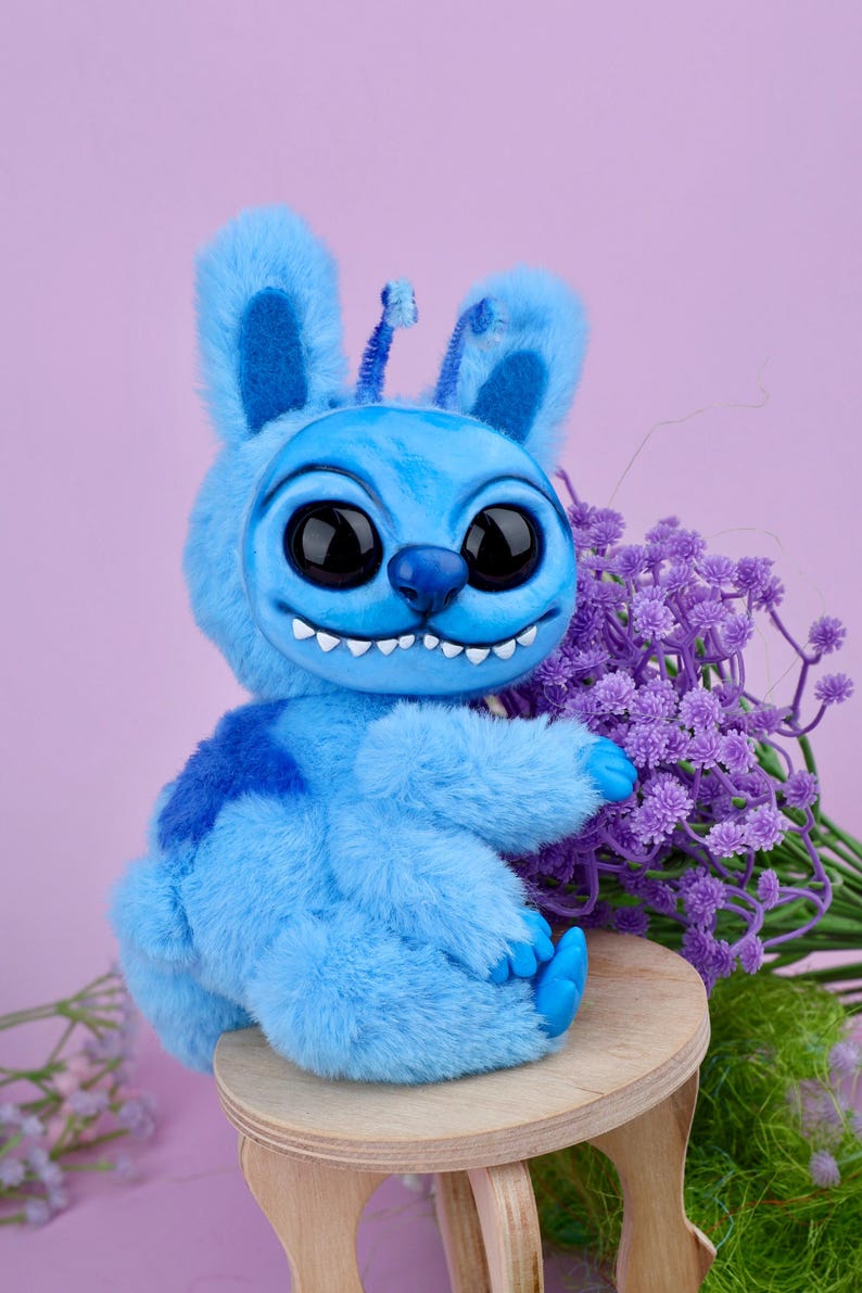 May include: A plush blue creature with large black eyes, a wide grin, and bunny-like ears. The stuffed animal is sitting on a small wooden stool, with a bouquet of purple flowers in the background. The toy has a patch of darker blue on its back.