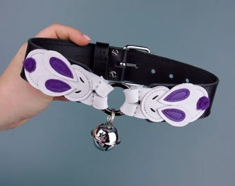 May include: Black leather collar with white and purple accents. The collar features a silver bell and decorative white shapes with purple inserts. The collar has a silver buckle and a D-ring.
