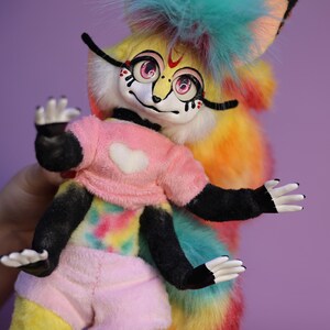 Op de afbeelding: Een kleurrijk pluche speelgoed met een regenboog tie-dye patroon, zwarte ledematen en een roze shirt met een wit hart. Het speelgoed heeft een pluche, turquoise blauwe mohawk en een lange, regenboogstaart.