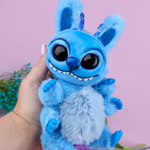 May include: A plush blue creature with large black eyes, a toothy grin, and fluffy fur. The toy has large ears and small arms and legs. The background is a solid pink color.