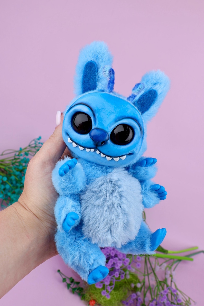 May include: A plush blue creature with large black eyes, a toothy grin, and fluffy fur. The toy has large ears and small arms and legs. The background is a solid pink color.