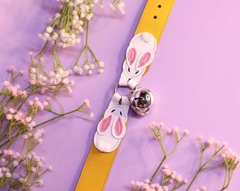 May include: A yellow leather collar with two white bunny appliques and a silver bell. The collar is on a purple background with white flowers.