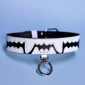 May include: A white leather collar with black bat-shaped cutouts and a silver ring attached. The collar is stitched with white thread.