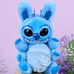May include: A blue plush toy resembling a cartoon character, with large black eyes, a wide grin, and fluffy fur. The figure is seated on a small wooden stool, with a light purple background and some purple flowers.