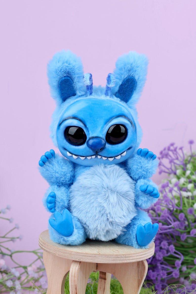 May include: A blue plush toy resembling a cartoon character, with large black eyes, a wide grin, and fluffy fur. The figure is seated on a small wooden stool, with a light purple background and some purple flowers.