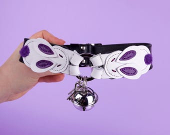 May include: A black leather collar with white and purple bunny-shaped accents. The collar features a silver bell and key charms. The bunny shapes have purple accents and a small purple pom-pom. The background is a solid purple.