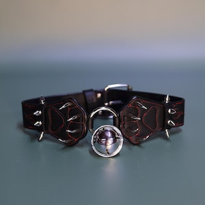 Puede incluir: Collar de cuero negro con costuras rojas y huellas de patas plateadas. El collar tiene una campana plateada.