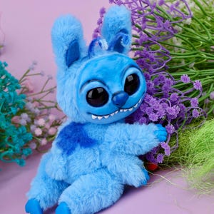 May include: A plush blue creature with large black eyes, a mischievous grin, and fluffy fur. The toy has a darker blue patch on its chest and is posed with its arms outstretched. The background features purple and green floral accents against a pink backdrop.