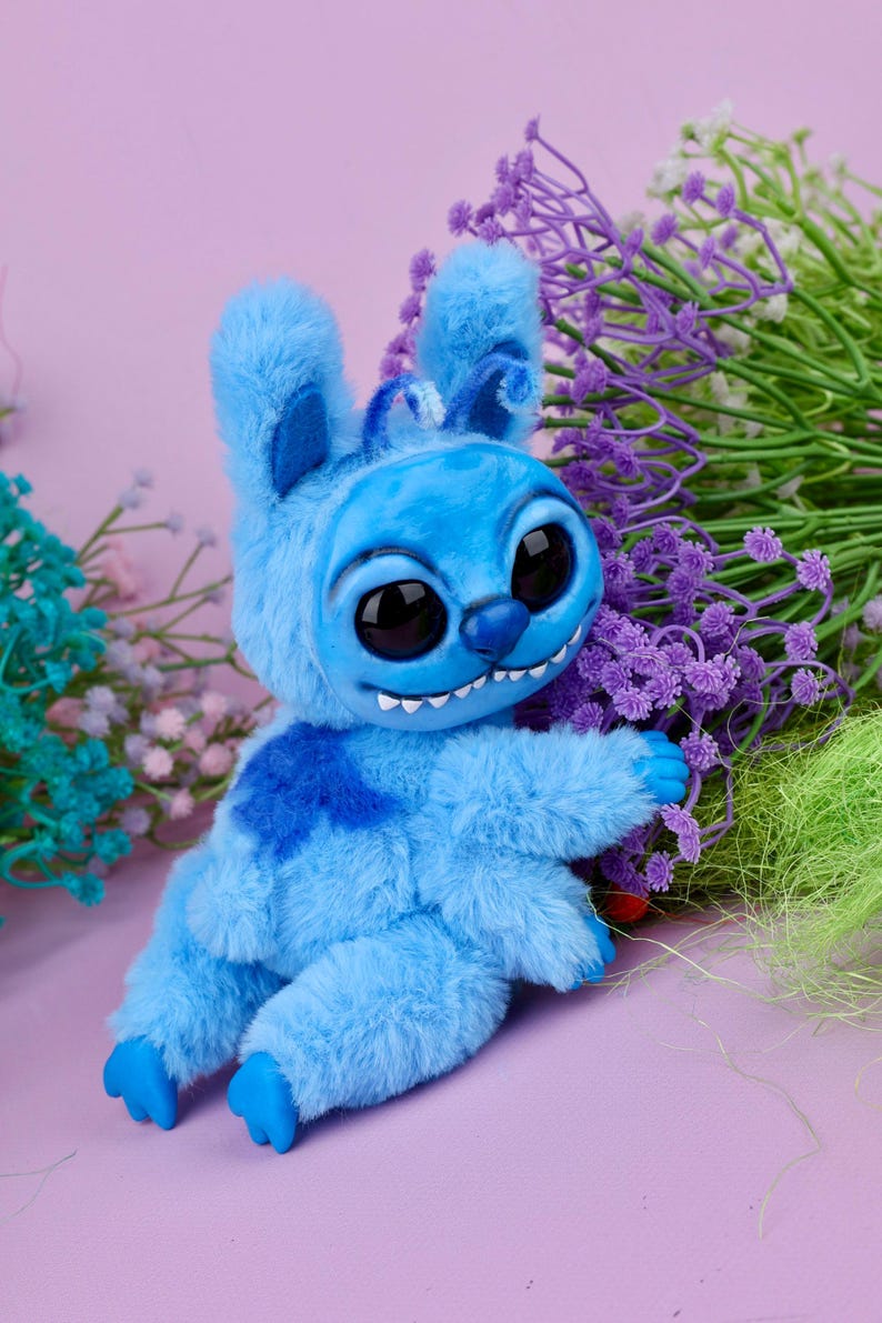May include: A plush blue creature with large black eyes, a mischievous grin, and fluffy fur. The toy has a darker blue patch on its chest and is posed with its arms outstretched. The background features purple and green floral accents against a pink backdrop.