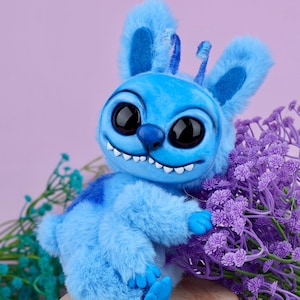 May include: A blue plush toy with large black eyes, a toothy grin, and fluffy ears. The toy is sitting on a small wooden stool next to purple flowers. The background is a soft purple.