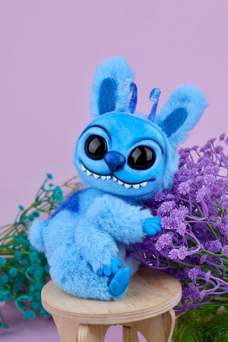 May include: A blue plush toy with large black eyes, a toothy grin, and fluffy ears. The toy is sitting on a small wooden stool next to purple flowers. The background is a soft purple.