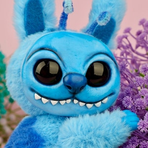 May include: A plush, blue creature with large, black eyes and a wide grin. The handmade toy has large ears, a tufted body, and a small antenna. The background is a soft pink with purple flowers.