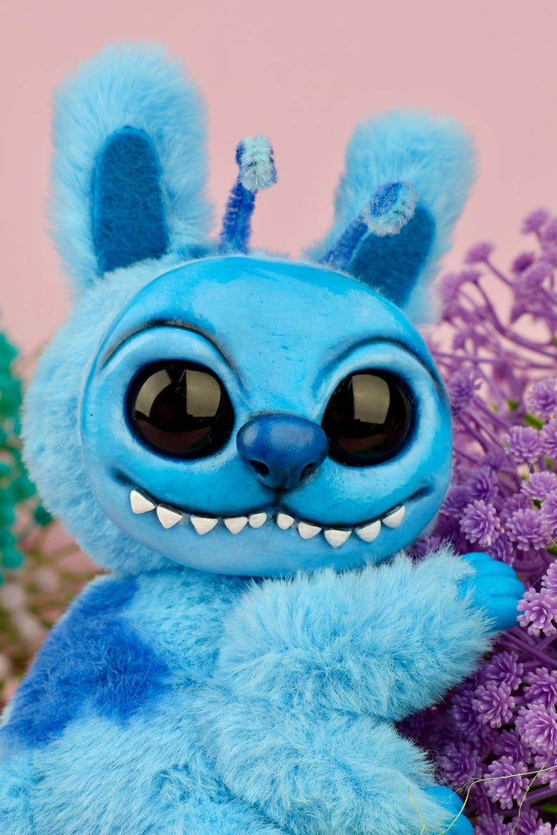 May include: A plush, blue creature with large, black eyes and a wide grin. The handmade toy has large ears, a tufted body, and a small antenna. The background is a soft pink with purple flowers.