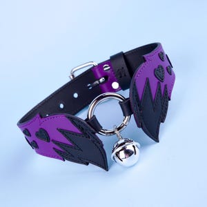 May include: A black and purple collar with a silver bell. The collar features purple wing-shaped accents with black detailing and heart-shaped cutouts. The collar has a silver metal ring and buckle.