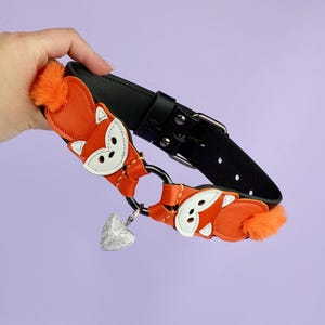 May include: A black leather collar featuring orange fox-shaped details and a silver heart charm. The fox designs have white faces and orange fur tails. The collar has a metal buckle and a D-ring.