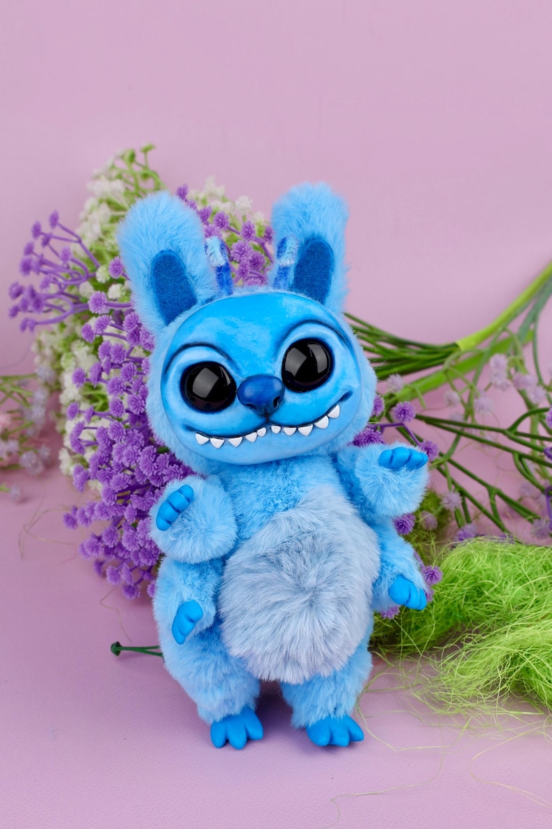 May include: A plush blue creature with large black eyes, a wide grin, and fluffy ears. The toy has a light blue belly and blue feet and hands. It is posed in front of a bouquet of purple and white flowers.