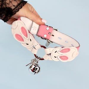 May include: A white and pink leather collar with bunny ear accents and a silver bell charm. The collar features a pink leather band and a silver buckle. The bunny ears have pink inner ears and white fur. The bell is silver and has star cutouts.