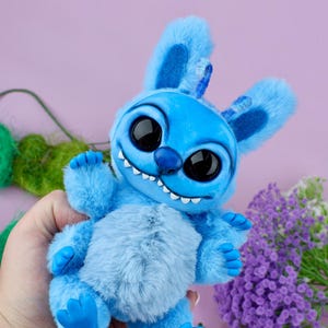 May include: A plush blue creature with large black eyes, a wide grin, and fluffy fur. The toy has large ears and small blue feet. The background is a soft purple with some green and purple floral accents.