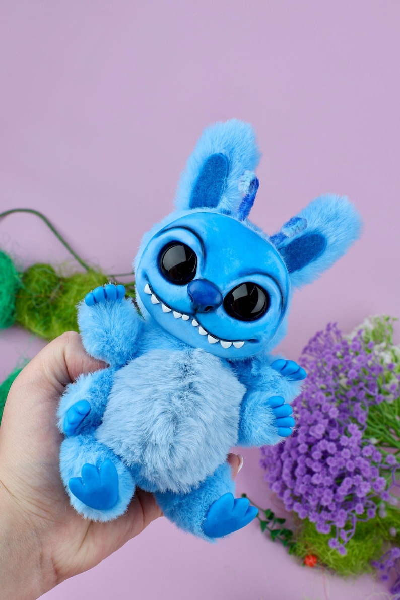 May include: A plush blue creature with large black eyes, a wide grin, and fluffy fur. The toy has large ears and small blue feet. The background is a soft purple with some green and purple floral accents.