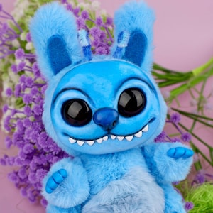 May include: A blue plush creature with large black eyes, a wide grin, and fluffy ears. The toy has a light blue belly and blue hands. The background features purple flowers and a pink backdrop, creating a whimsical and playful aesthetic.