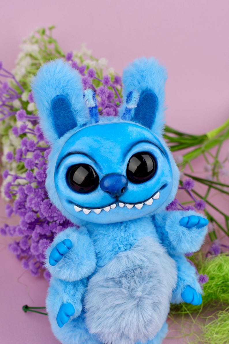 May include: A blue plush creature with large black eyes, a wide grin, and fluffy ears. The toy has a light blue belly and blue hands. The background features purple flowers and a pink backdrop, creating a whimsical and playful aesthetic.