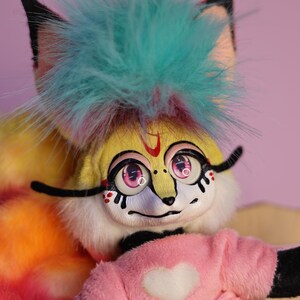 Op de afbeelding: Een geel en wit pluche speelgoed met een turquoise mohawk, zwarte ogen, roze oogleden en een roze hart op een roze shirt. Het speelgoed heeft een tie-dye staart.