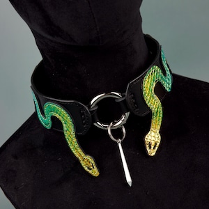 May include: A black leather choker featuring two iridescent green and yellow snake appliqués on either side. The centre of the collar includes a large silver-toned metal O-ring with a smaller dangling silver-toned spike charm attached to it.