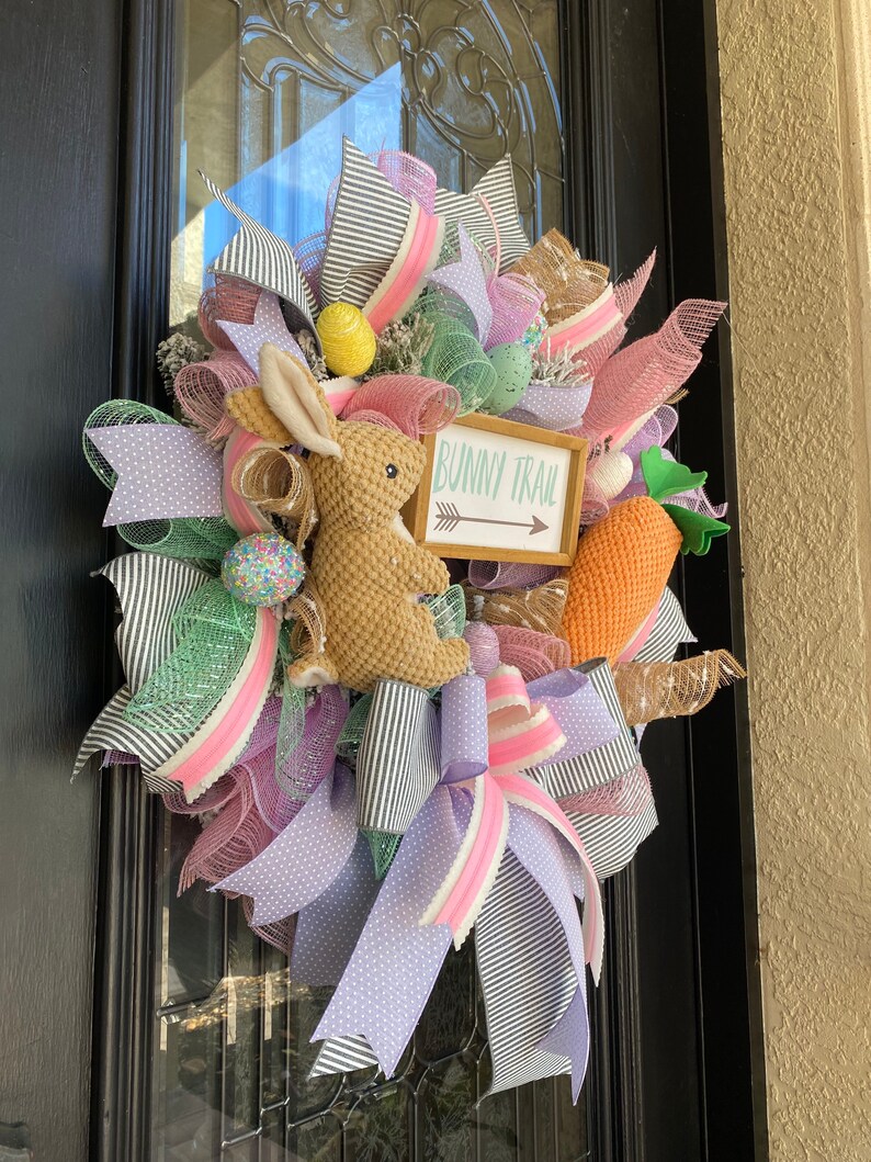 May include: Easter wreath with a woven bunny, decorative eggs, and a carrot. The wreath features ribbons in pink, lavender, green, and black and white stripes. A sign reads "Bunny Trail" with an arrow.