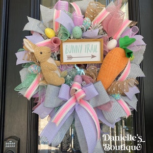 May include: A decorative Easter wreath with a bunny, carrot, and eggs. The wreath features a sign that reads "Bunny Trail" with an arrow. The wreath is decorated with pink, purple, and white ribbons, and is displayed on a black door.