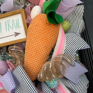 May include: Easter wreath with a large orange carrot, green leaves, and a sign that says "Easter Trail". The wreath features pink, purple, and white ribbons, along with decorative eggs and a textured carrot.