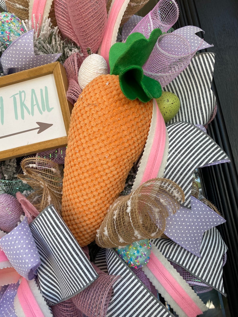 May include: Easter wreath with a large orange carrot, green leaves, and a sign that says "Easter Trail". The wreath features pink, purple, and white ribbons, along with decorative eggs and a textured carrot.